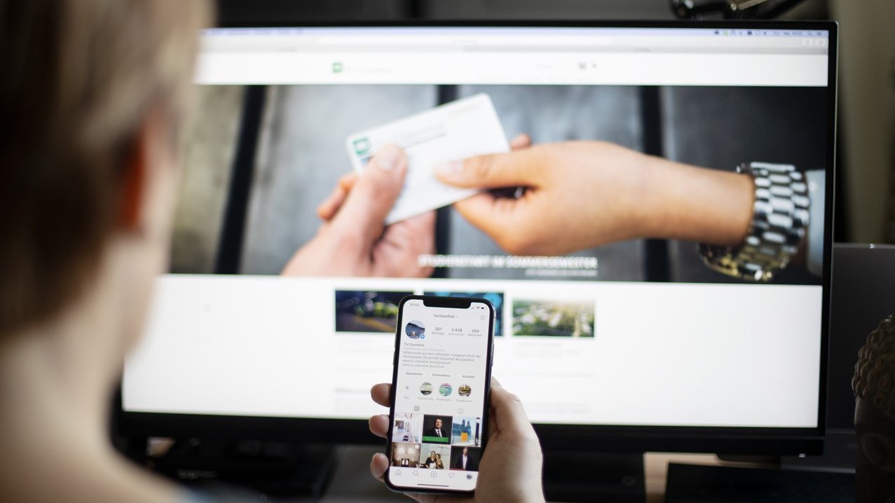 Person sitzt mit Handy vor einem Bildschirm